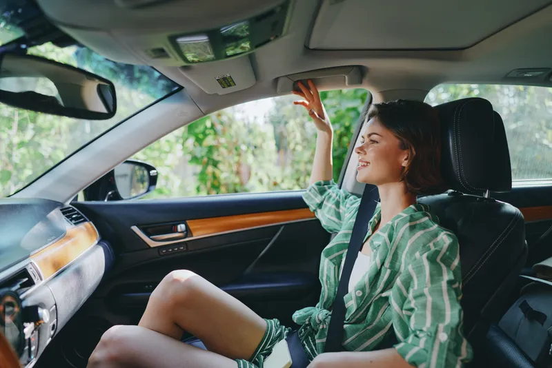 A stylish woman sits in a car seat, smiling and waving, enjoying a sunny drive inside a sleek interior with wood trim.