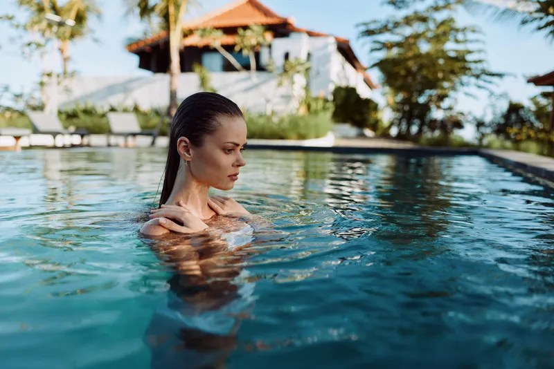 A young woman with wet hair stands in a clear outdoor pool near a tropical villa during daytime. The lifestyle scene conveys relaxation and peacefulness in a sunny environment.