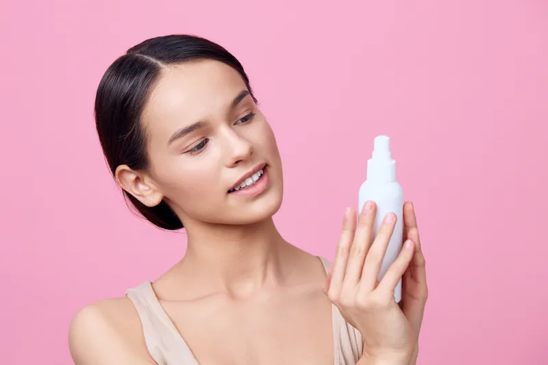 Young woman examining a skincare spray bottle, showcasing a fresh and natural beauty concept with a pink background