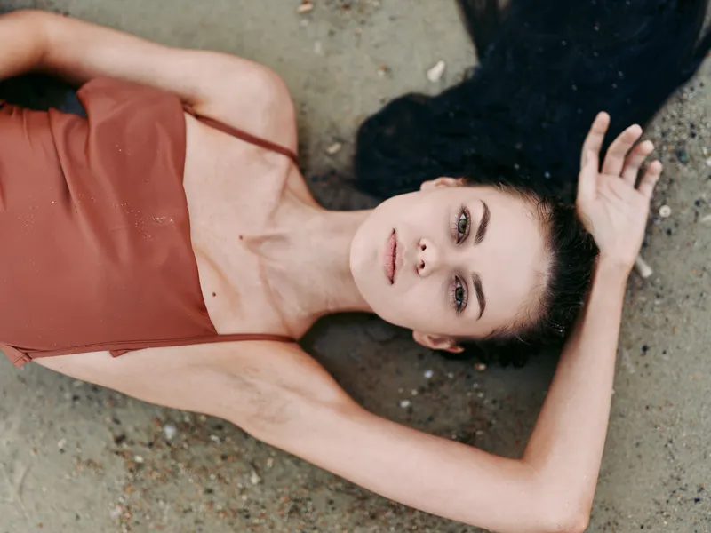 A female model lies on a sandy surface, wearing a rust colored dress with thin straps, gazing softly at the camera, in a relaxed, sunlit pose.