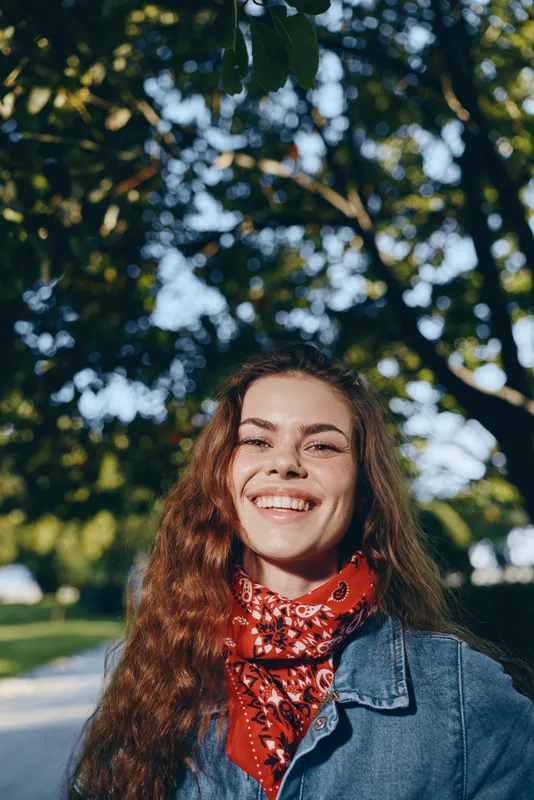 Smiling woman in modern boho-western denim street style with a vibrant red bandana, natural outdoor setting, and warm film color tones conveying lifestyle and joy.