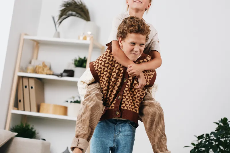 Two boys share a playful piggyback moment indoors, wearing knit sweaters and denim as they laugh and balance, capturing a lively, heartwarming scene of youthful friendship.