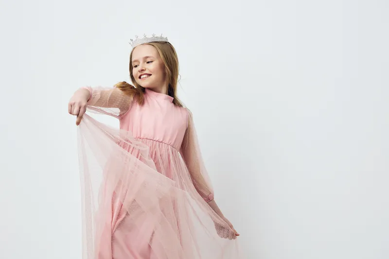 Joyful girl in a pink tulle dress, wearing a crown, showcasing playful emotions against a simple light background, perfect for children s fashion or celebration themes