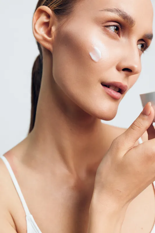 Closeup of a woman applying cream on her cheek, showcasing skincare routine, smooth skin, gentle glow, minimal makeup, calm expression, soft lighting, intimate portrait