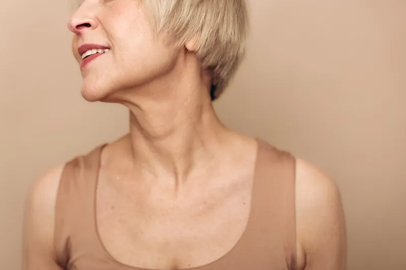 Closeup of mature woman with short blonde hair looking sideways in beige top against neutral background. Concept for aging skin, natural beauty, and confident lifestyle.