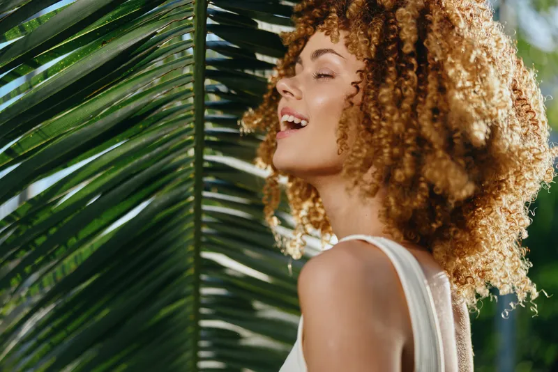Smiling woman with curly hair enjoys sunlight near green tropical leaves, wearing white sleeveless top in bright outdoor natural setting, vibrant and joyful moment.