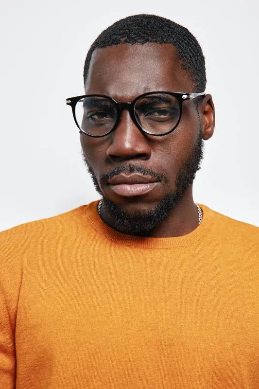 Portrait of African American man with dark skin wearing glasses and orange sweater looking serious on white background. Closeup, facial expression and casual style with short hair and beard.