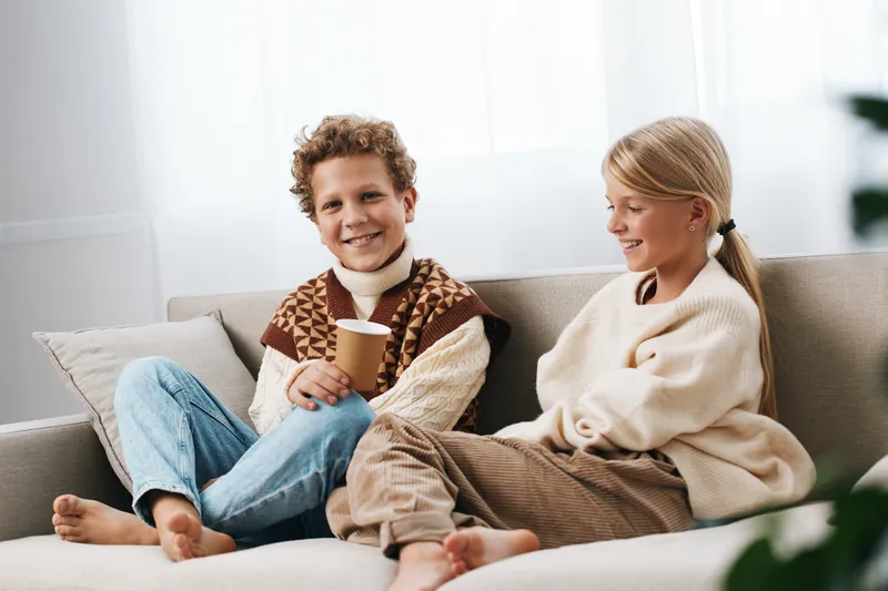 Two children sit on a cozy sofa indoors, smiling and relaxed as one holds a warm cup while wearing knit sweaters. Soft natural light captures a simple, candid moment of comfort.