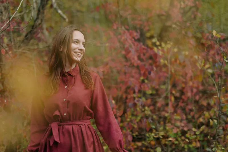 An authentic moment features a smiling woman in a deep burgundy dress standing in a sunlit autumn forest, conveying honest charm, calm presence, and natural beauty in soft light.