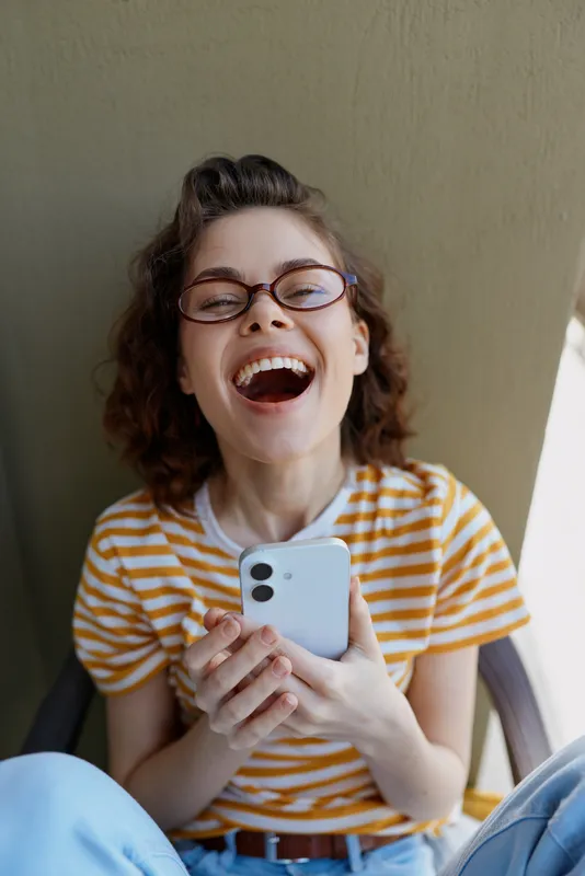 Cheerful woman indoors, laughing with mouth open, striped shirt, glasses, holding smartphone, casual lifestyle moment, warm light, relaxed pose, natural colors, cozy home setting, ambience
