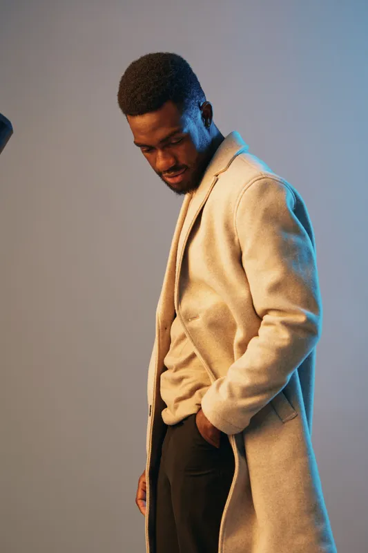 Stylish young man in a beige coat and sweater displaying confidence in a creative studio setting with a muted background