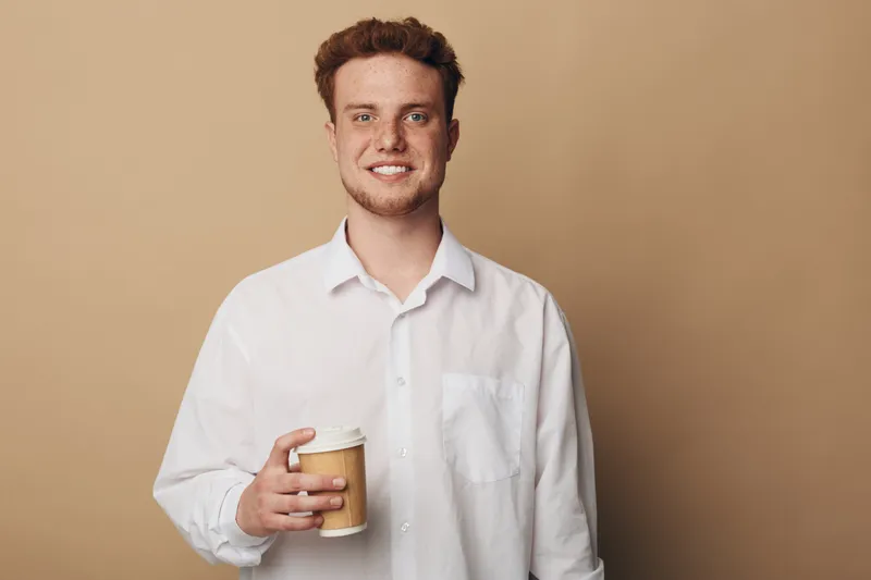Smiling young man in white shirt holds disposable coffee cup against beige background. Casual lifestyle concept with positive mood and relaxed expression in studio portrait.