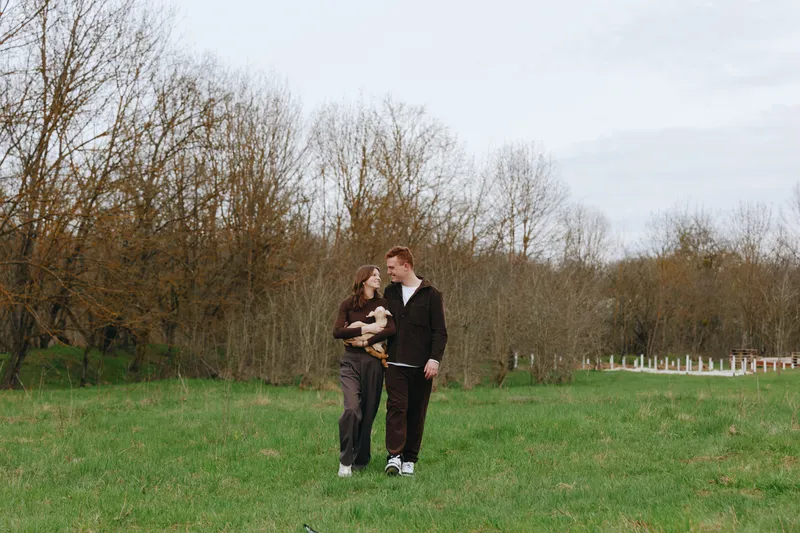 Cheerful couple outdoors, walking in a grassy park, one person holding a small dog, autumn trees in the background, relaxed mood, natural colors, candid moment, gentle breeze, lifestyle scene