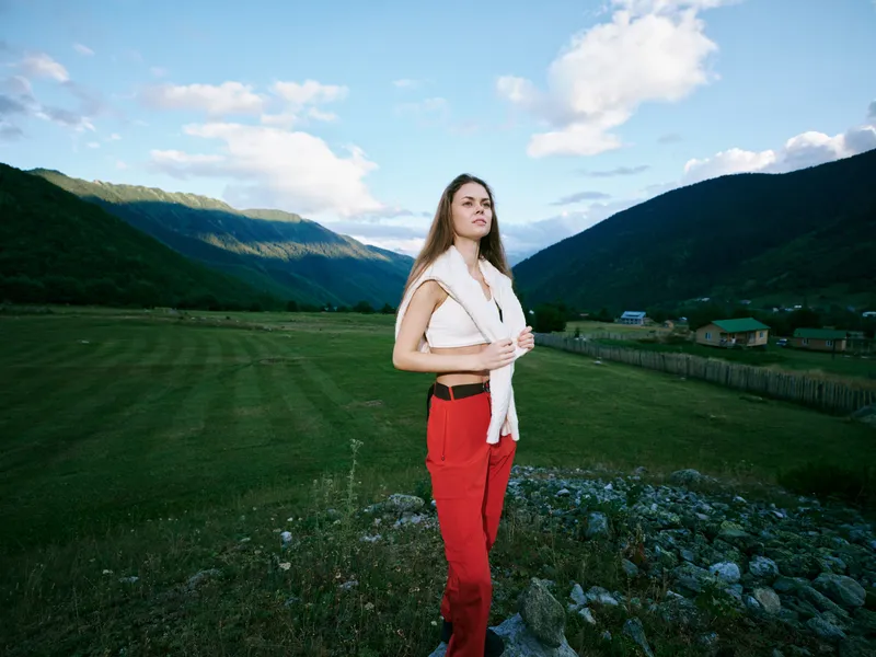 Woman standing in a wide valley with green fields and distant mountains, casual red pants and white scarf, sunny day, blue sky, open landscape, exploration and freedom in nature