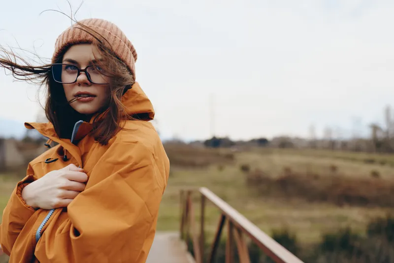 Lifestyle woman outdoors in autumn coat and knit hat, wearing glasses, standing on rural bridge walkway, wind blowing hair, calm thoughtful expression, bright soft daylight