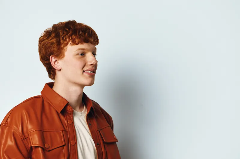 Young man with red hair and freckles wearing a brown leather jacket and white t shirt standing smiling against a light background in profile view.