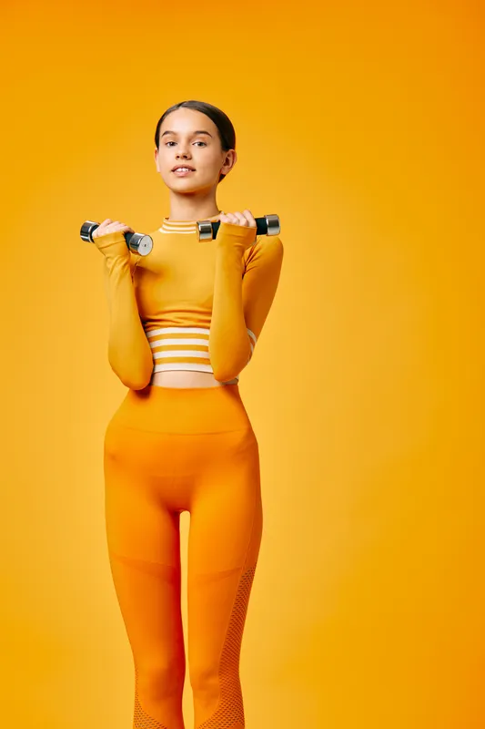 Fitness portrait of a confident woman in orange athletic wear lifting dumbbells in a bright studio, showcasing strength, balance and energy against a vivid orange backdrop.