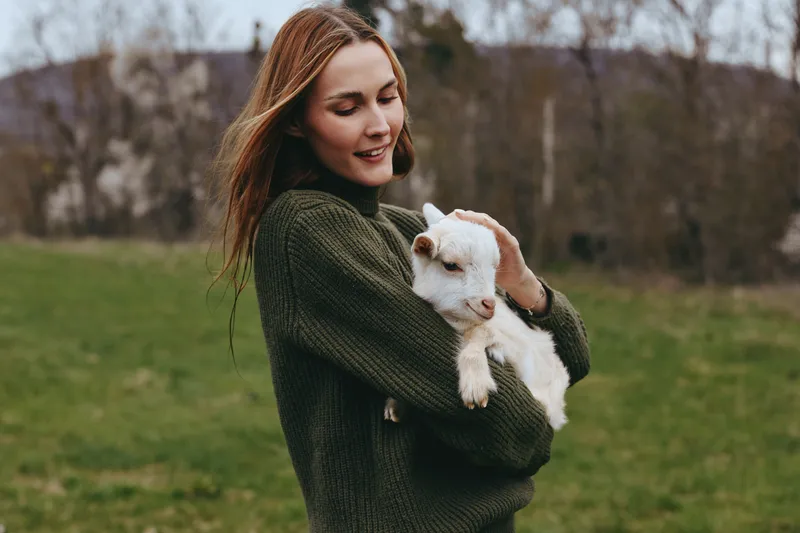 Smiling woman in a dark sweater holds a goat kid outdoors, capturing lifestyle, countryside scenery, affection, and connection with nature in a warm portrait. Bright natural light enhances mood.
