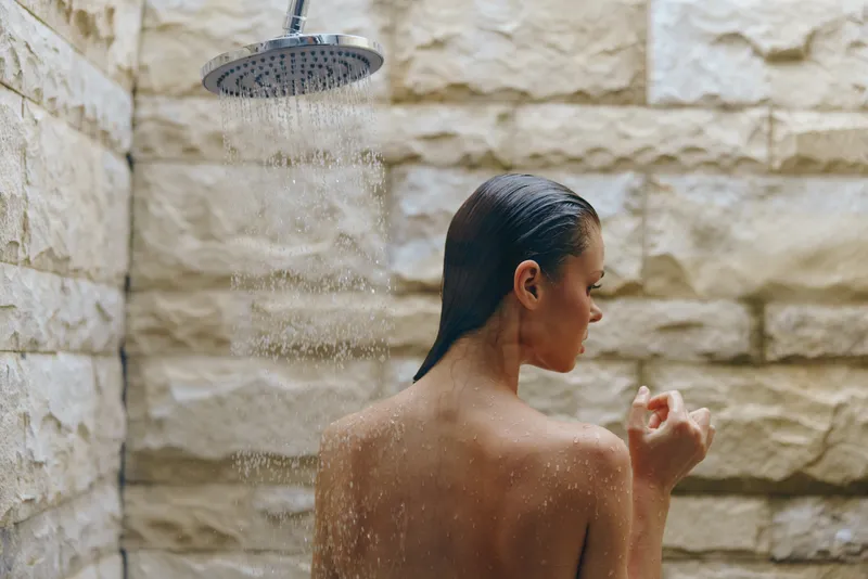 young woman taking a shower under rain showerhead, wet hair, natural stone wall background, relaxing moment in bathroom