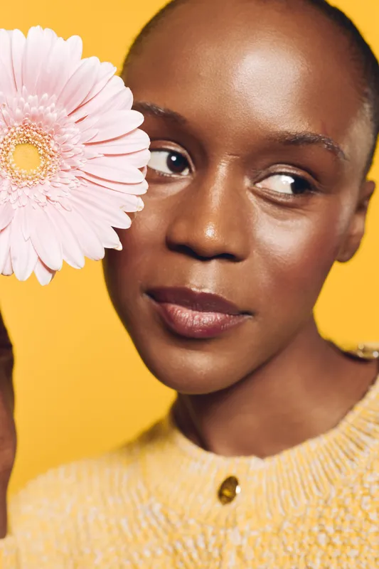 Close-up of an African woman with dark skin holding a pink flower near her face. She wears a yellow sweater and looks to the side against a bright yellow background. Concept of beauty and nature.