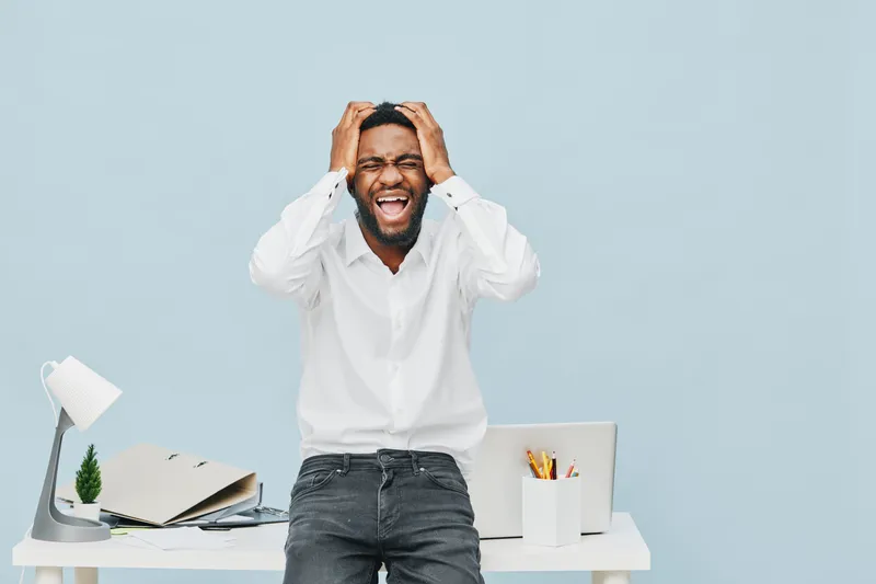 Joyful excited young man reacting with surprise and happiness while standing at his modern home office desk, holding his head with both hands, wearing casual white shirt and jeans, cheerful facial exp