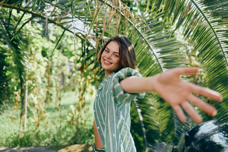 Cheerful woman smiles and reaches toward the camera in a lush sunlit tropical garden, capturing a carefree and vibrant moment.