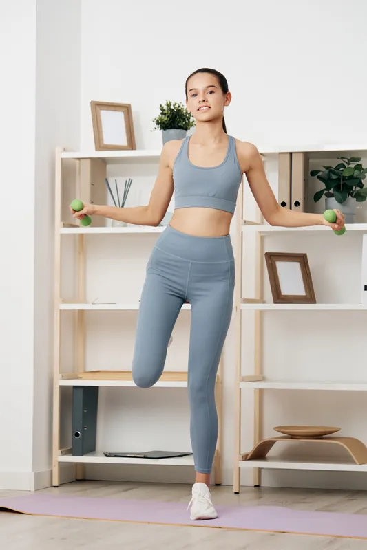 A woman in blue athletic wear performs a balance exercise indoors, lifting small dumbbells beside a neat home gym setup with shelves, plants and frames in bright, minimal space.