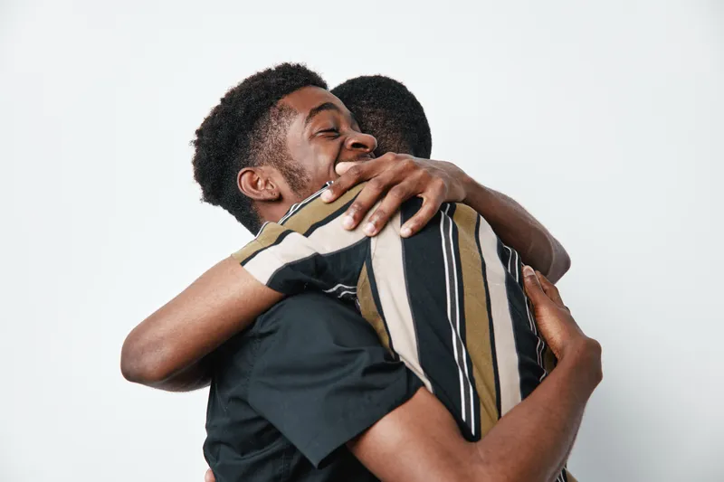 Two young black men hugging each other with happy expressions on white background. Friendship, connection and support between african american males in casual clothing.