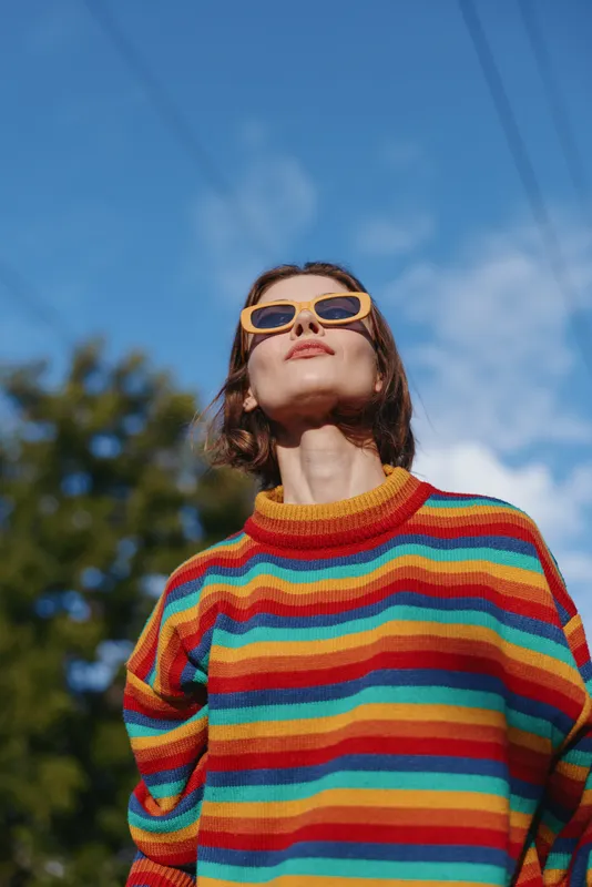 Young adult woman in a colorful striped sweater and yellow sunglasses looking up outdoors against a blue sky, casual outfit, confident pose, lifestyle and fashion portrait.