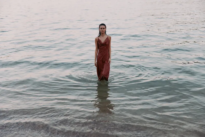 A solitary woman stands waist deep in calm sea, wearing a rust red dress that clings to her form as gentle waves lap the shore and warm light glows on the water.