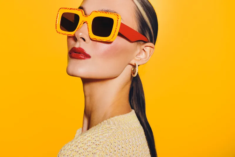 Stylish woman with slicked back hair wears oversized orange sunglasses and red lipstick against bright yellow background. Concept of fashion, beauty, and summer style.