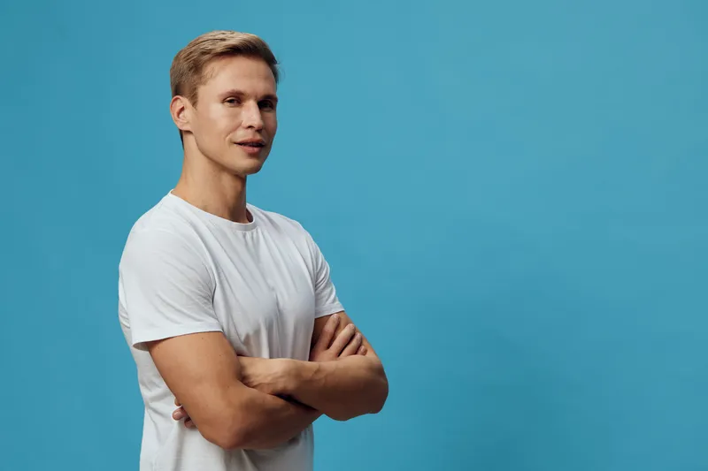 Male young adult with casual white t-shirt posing confidently against a plain blue background. Studio portrait emphasizing relaxed attitude, modern style, and natural appearance. Concept: casual confi