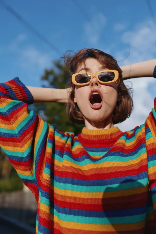 Young adult woman in a rainbow sweater and yellow sunglasses outdoors, short hair with surprised expression, hands behind head, casual outfit and bright sunny lifestyle portrait.