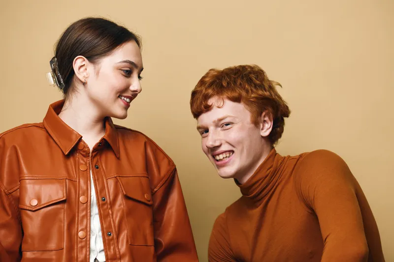 Two young people with red and brown hair wearing brown clothing smiling and interacting on beige background in casual indoor portrait studio lighting.