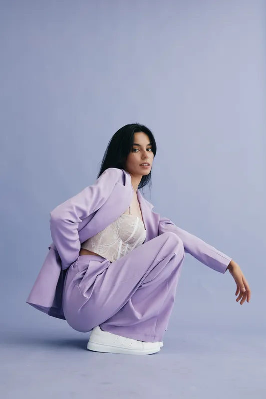 Woman in lilac suit and white sneakers crouching confidently against plain light blue background, showcasing modern fashion and casual style.