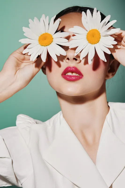 Creative close-up of woman with white daisies covering eyes, wearing glossy pink lipstick and a white outfit. Bright colorful makeup concept for beauty and fashion editorial.