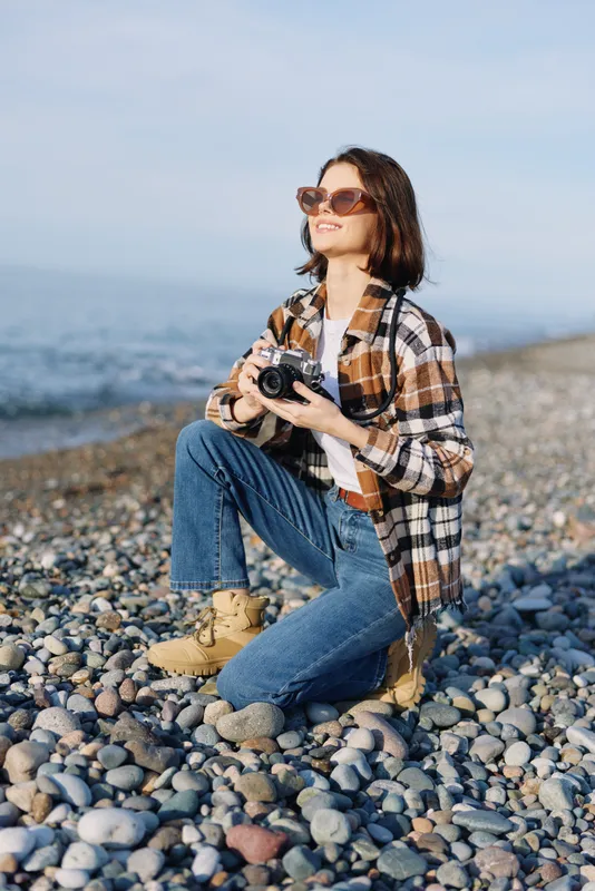 A woman photographer sits on a rocky beach, capturing scenery with a handheld camera, wearing sunglasses, a plaid shirt and jeans in daylight.