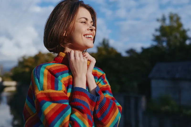 Woman smiling in a colorful rainbow sweater and short hair outdoors, young adult enjoying autumn sunlight; casual outfit, hands near collar, joyful lifestyle and travel mood.