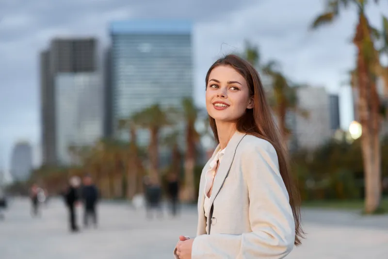 Young woman smiling confidently outdoors in beige blazer with urban cityscape and palm trees in background during daylight KEY: woman confident city urban stylish blazer outdoor palm trees young natur