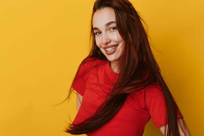 Young woman with long hair smiling and wearing red t shirt posing against yellow background. Portrait of happy female with natural makeup and casual style in studio lighting.