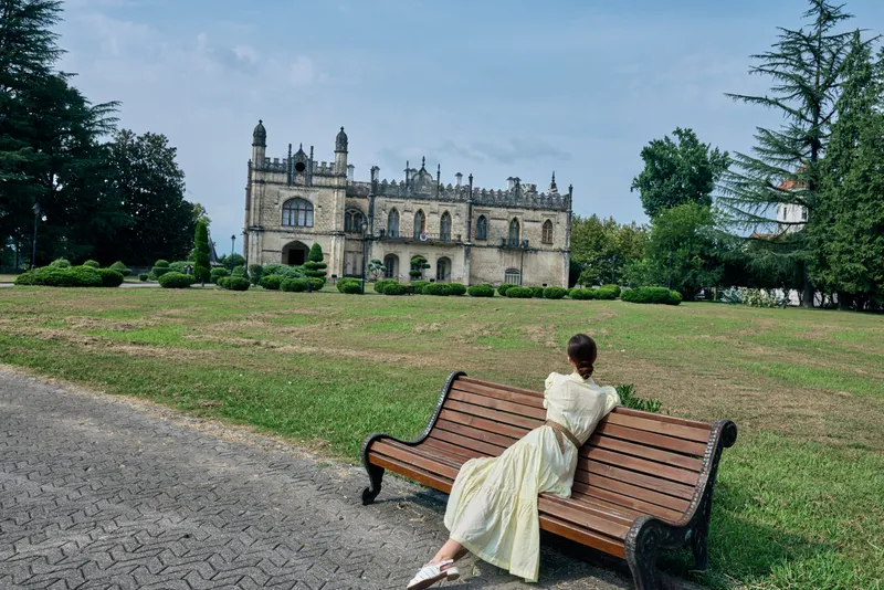 woman in a flowing dress sits on a bench in a park with a historic castle in the background, a peaceful outdoor landscape scene that conveys elegance and leisure