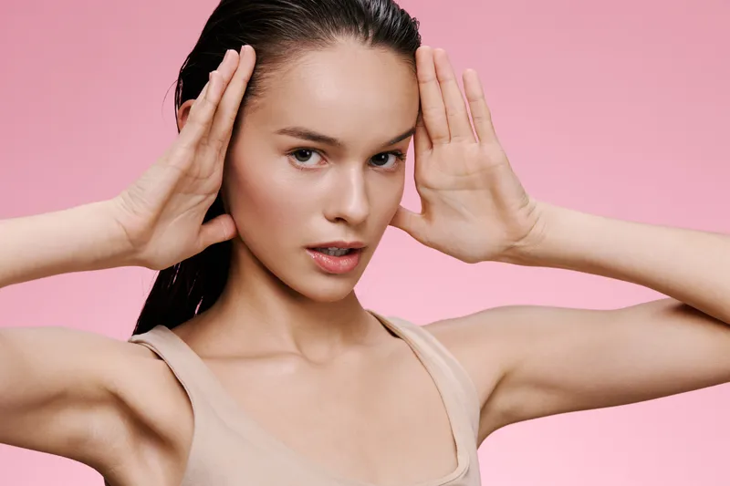 Young model with long hair posing against a soft pink background, expressing confidence and elegance in a light beige top, ideal for beauty and fashion concepts