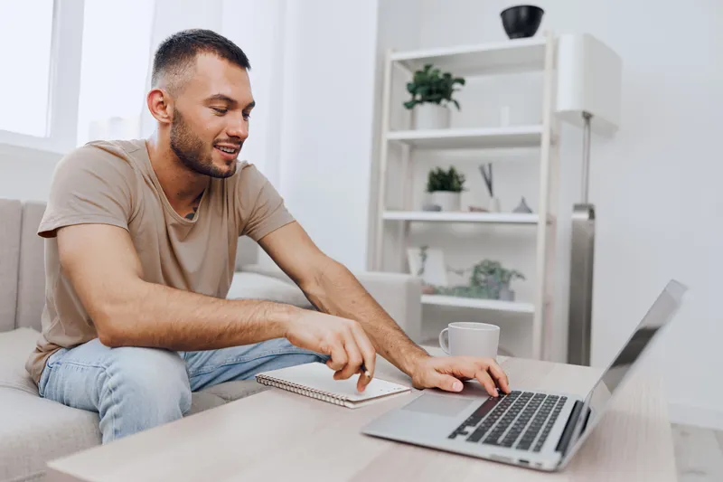 Smiling man working remotely on laptop, casual attire, modern home office, engaged in creative task, showcasing productivity and comfort in a relaxed atmosphere.