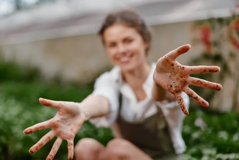 A joyful woman in a garden with dirt covered hands, showcasing her love for gardening and nature, wearing casual clothing and smiling brightly amidst green foliage