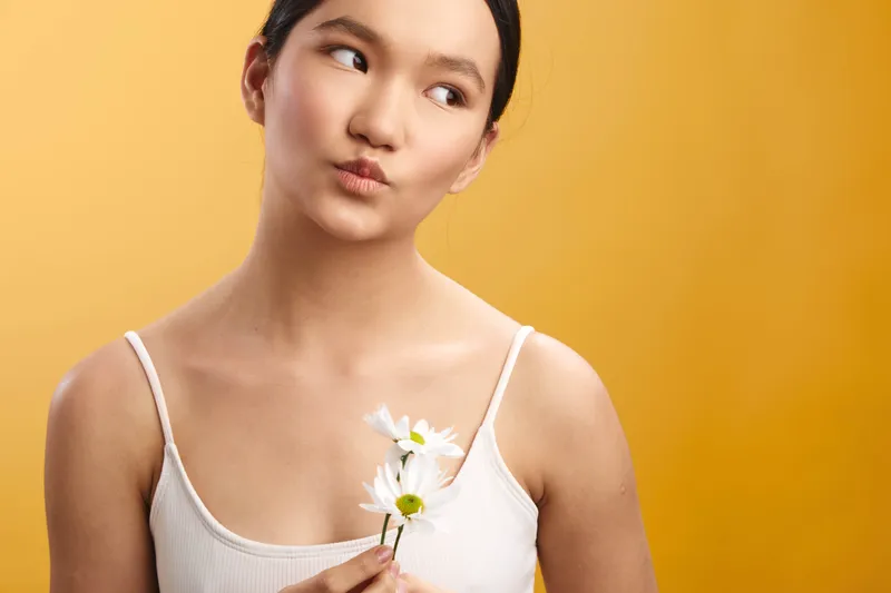 asian woman in a white tank top holds a small flower against a warm orange background, creating a calm, thoughtful portrait with soft light and a gentle, minimalist mood.