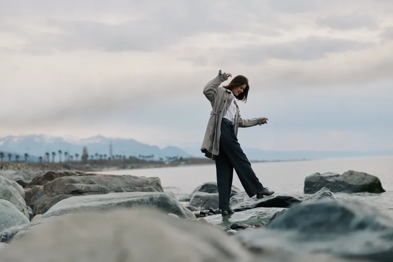 Young woman in casual clothes balances on rocks by the sea during cloudy weather. Concept of leisure, nature, and outdoor activity near water and mountains.
