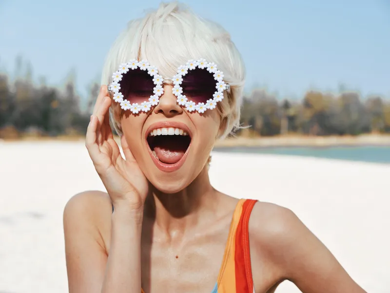 Cheerful woman on a sunlit beach wears oversized circular flower sunglasses, laughing with mouth open, capturing bold playful fashion and carefree summer vibe.