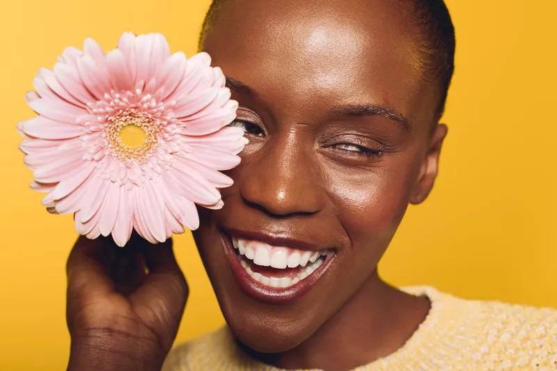 Happy African woman with dark skin smiles joyfully holding pink gerbera flower near face, wearing yellow sweater on bright yellow background. Concept of beauty, skincare, and positive mood.
