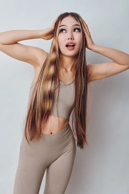 Young woman with long straight hair wearing beige sportswear posing with hands on head looking surprised against light background.