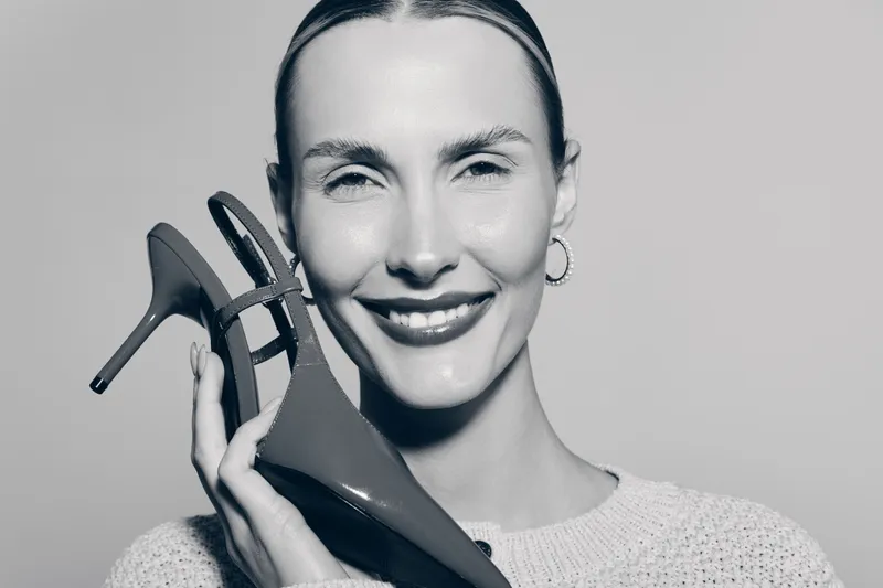 A smiling white woman with fair skin holds a high heel shoe near her face, wearing a light sweater and hoop earrings on a plain background. Concept of fashion, beauty, and style.